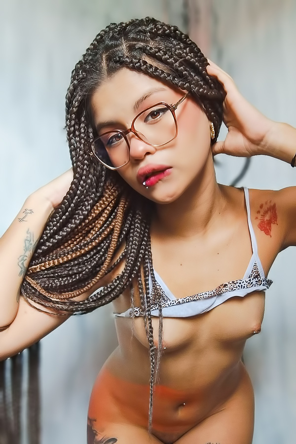 Lilith Baph in white bikini poses with leg up showcasing her tattooed curves and braids indoors
