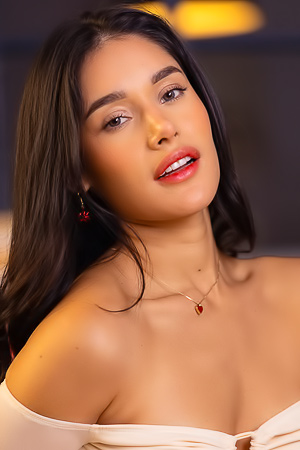 Carolina Guerrero teases in denim shorts and white off shoulder top, legs spread on bed, red earrings gleaming