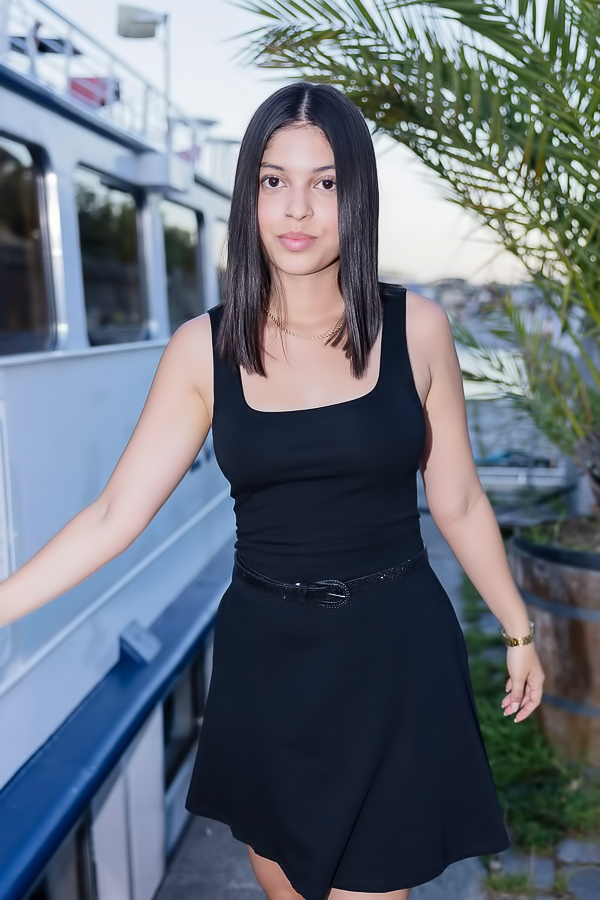Gia Rosse leans against stone wall in black dress outdoors posing with gold necklace and wristwatch
